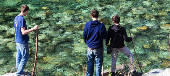 teenagers standing on the edge of a shallow, clear, wide river and looking into the water