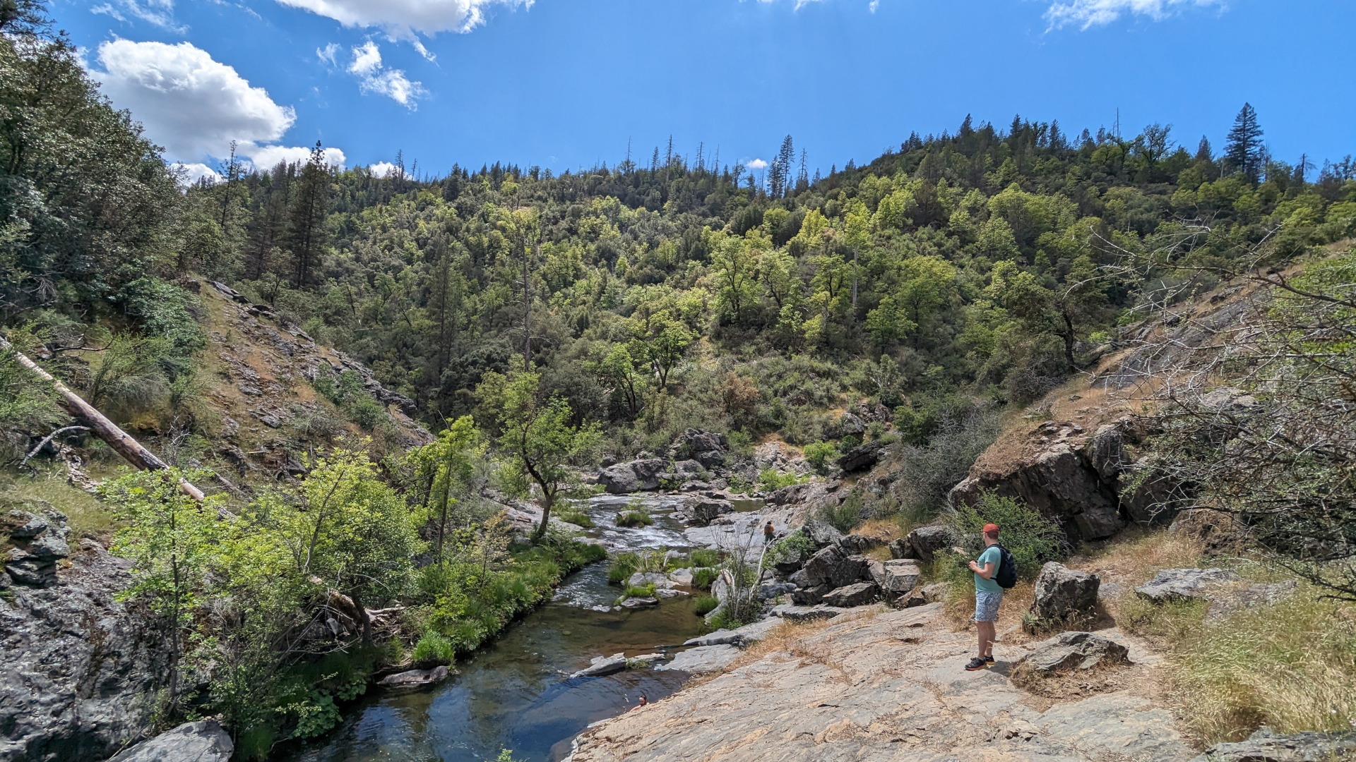 A man taking a break on a hike to stand on the rocky banks of a small river
